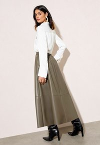 Pitkä oliivinvihreä tekonahkahame, jossa on poimutuksia, yhdistetty valkoiseen nappipaitaan. Mustat korolliset saappaat täydentävät asua.