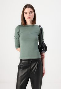 Grønn topp med tre kvarte ermer i et mykt materiale, sammen med svarte skinnbukser og en liten svart håndveske. Enkelt design og tettsittende fasong.