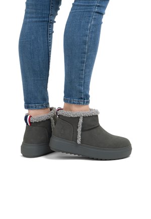 Henkilö, jolla on päällään harmaat talvijalanpohjaiset saappaat, joissa on fleece-vuori ja reunus, yhdistettynä sinisiin skinny-farkkuihin, seisoo valkoista taustaa vasten.