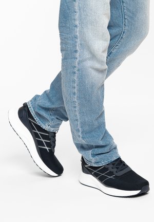 Persona con jeans azul claro y zapatillas deportivas negras con suelas blancas, un pie ligeramente levantado como si estuviera caminando.