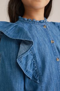 Denim srajca z nabranimi rokavi, modre barve, okrogla izreza z nabranimi detajli in rjavimi gumbi. Gladka tekstura in sproščen oblikovni slog.