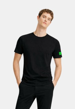 Giovane uomo con capelli corti e ricci indossa una maglietta nera semplice con una piccola toppa verde sulla manica sinistra, mani nelle tasche, sfondo neutro.