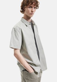 Camisa de botones de manga corta de color verde claro con un corte relajado, que presenta una textura suave y un dobladillo recto, llevada sobre una camiseta interior de color más oscuro.