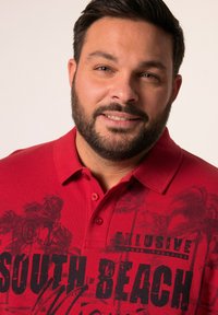 Camisa polo roja con cuello, que presenta un estampado gráfico negro de palmeras y el texto "SOUTH BEACH" en el frente. Dos botones en el cuello.
