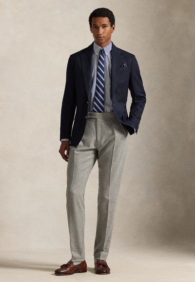 Blazer azul-marinho, camisa às riscas azul e branco, calças cinzentas plissadas, sapatos de couro castanhos com detalhes e um lenço de bolso.