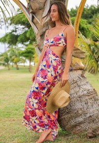 Robe maxi avec un motif floral en rose, orange et bleu, présentant un détail noué, des côtés découpés et un bas fluide. Tenant un chapeau en paille.