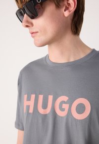 Pilkas medvilnės marškinėlis su dideliu rožiniu "HUGO" logotipo spaudiniu. Dizainas turi apvalų kaklą ir trumpas rankoves, demonstruojant glotnią tekstūrą.
