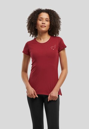 Femme aux cheveux bouclés portant un t-shirt rouge ajusté avec un petit contour blanc de visage de chien sur la poitrine et des leggings noirs sur fond clair.