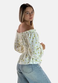 Weiße Bluse mit Carmenausschnitt und gelbem Blumenmuster, mit gerafftem Taillenbund und Puffärmeln; kombiniert mit hellblauen Jeans.