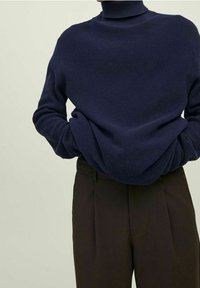 Persona che indossa un maglione a collo alto testurizzato blu navy, con le mani infilate nelle tasche di pantaloni a vita alta marroni scuri.