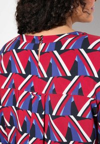 Rode top met geometrische blauwe en witte patronen. Voorzien van een knoopsluiting aan de achterkant en een kleine uitsnijding. Gemaakt van lichtgewicht stof.
