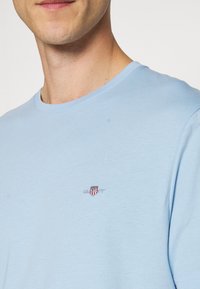 Hellblaues Baumwoll-T-Shirt mit Rundhalsausschnitt, kurzen Ärmeln und einem kleinen gestickten Logo mit einem Schild auf der linken Brust.