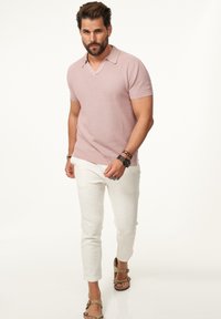 Kurzärmliges hellrosafarbenes Polohemd mit V-Ausschnitt, kombiniert mit weißen strukturierten Hosen und braunen Sandalen. Zubehör sind Armbänder.