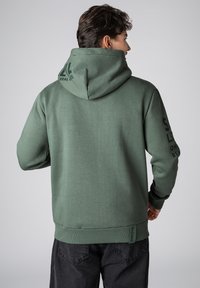 Groene hoodie met een textuur, voorzien van een capuchon en bedrukt accent op de mouwen. Ribbels aan de manchetten en onderrand met een merklabel aan de onderkant.