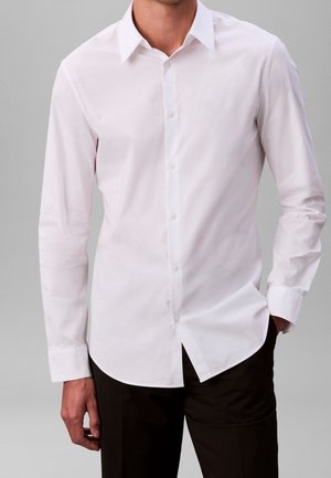 Hombre con camisa blanca de manga larga y cuello abotonada, combinada con pantalones oscuros sobre un fondo gris liso.