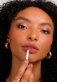 Kvinna med lockigt hår som applicerar mauve-läppglans, bärande guldhoopörhängen och en guldring, fokuserad på att noggrant applicera läppsmink.