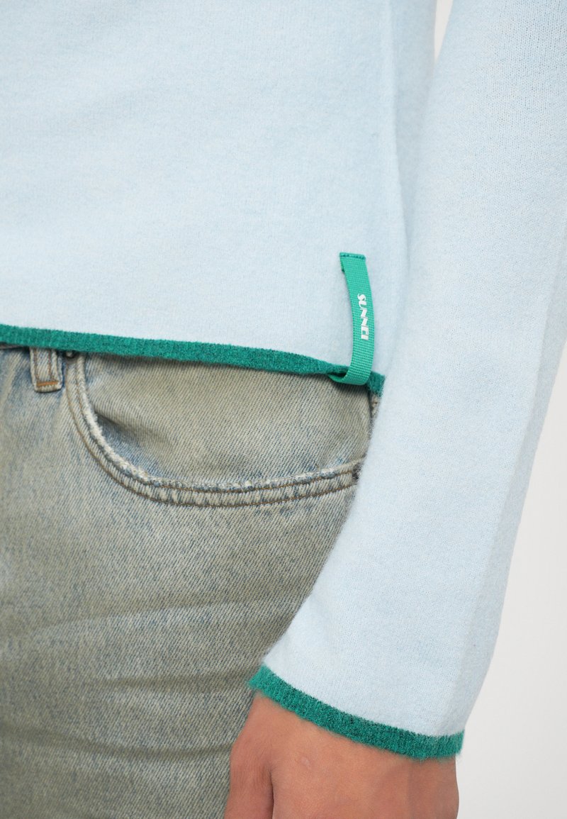 Ljust blått stickat tröja med gröna kantdetaljer. Har tagg i nedersta vänstra hörnet och en mjuk textur. Matchar med ljusa denimjeans.