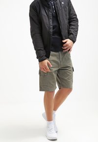 Una chaqueta bomber negra sobre un polo oscuro, combinada con pantalones cortos de cargo en color oliva. Zapatillas deportivas blancas completan el conjunto, con las mangas ligeramente arremangadas.