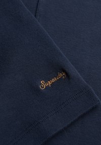 Navyblauwe katoenen stof met een gladde textuur, met een gouden "Superdry" geborduurd logo op de rand. Schoon ontwerp met subtiele stiksels.