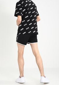 T-shirt noir avec des logos et du texte blancs répétés, associé à un short noir avec des bordures blanches, porté avec des baskets blanches.