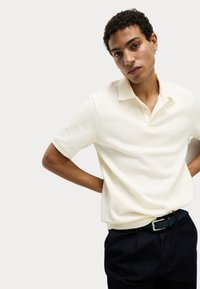Kortærmet polo shirt i blød, ribbet cremefarvet stof med krave og tre knapper, parret med mørke bukser og et sort bælte.