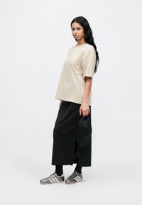 Bézs színű, túlméretezett pamut póló, fekete hosszú szoknya oldalsó hasítékkal, szürke-fehér sportcipő, és fekete sapkát tart.