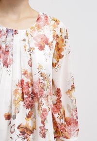 Blusa floreale con un tessuto leggero e trasparente. Presenta una base crema decorata con motivi di fiori rosa, arancioni e gialli. Collo plissettato.