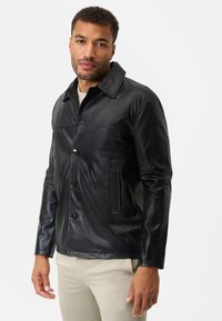 Schwarze Lederjacke mit einem knopfbedeckten Frontteil, großem Kragen, zwei Seitentaschen und einer glatten Textur. Kombiniert mit hellen Hosen.
