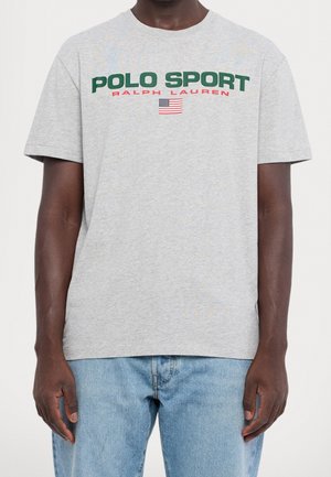 Szara koszulka z krótkim rękawem z "Polo Sport Ralph Lauren" w zielonym i czerwonym tekście nad małą amerykańską flagą, noszona z jasnoniebieskimi dżinsami.