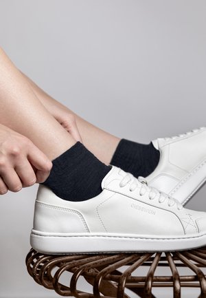 Białe skórzane sneakersy z teksturowanym wierzchem, okrągłym noskiem i płaskimi sznurowadłami. Widoczne czarne skarpetki za kostkę. Ustawione na zakrzywionej metalowej powierzchni.