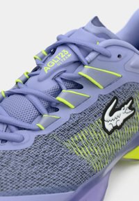 Violeta sporta apavi ar sietveida augšdaļu, neonzaļiem akcentiem un izteiksmīgu melnu Lacoste logo. Iespējas robustas auklas un teksturēti dizaina elementi.