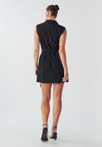 Kobieta w czarnej, bezrękawnikowej sukience-blazer z paskiem oraz czarnych, wysokich sandałach na obcasie, stojąca tyłem do aparatu.