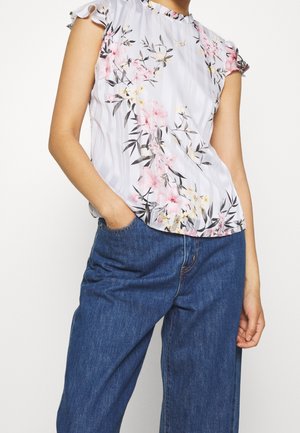 Vrouw draagt een witte korte mouwen bloemenblouse met ruches aan de mouwen en donkerblauwe hoog getailleerde spijkerbroek, hand in zak.