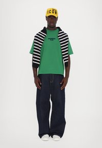 Grønn T-skjorte med kort erm og svart logo, oversized mørkeblå jeans, hvite joggesko og en gul caps med svart tekst, stripete genser over.