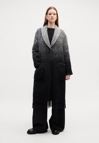 Cappotto lungo con gradiente dal grigio al nero, realizzato in tessuto testurizzato. Presenta un colletto a scialle, due tasche frontali e frange all'orlo.