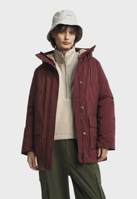 Bordeauxkleurige gewatteerde jas met capuchon, voorzien van twee voorkeisten, een lichtgewicht fleece binnenlaag en een ritssluiting. Draagt over olijfgroene broek.