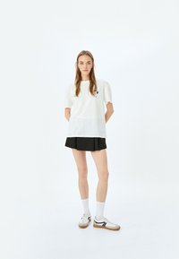 Witte t-shirt met korte mouwen en een grafische print op de linker borst, gecombineerd met een zwarte plissérok en witte sneakers met zwarte accenten.
