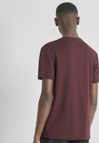 Hombre con cabello corto que lleva una camiseta de manga corta lila lisa, de pie de espaldas contra un fondo blanco liso.