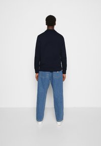 Homme debout de dos portant une veste bleu marine foncé, un jean large bleu clair et des chaussures blanches sur un fond uni clair.