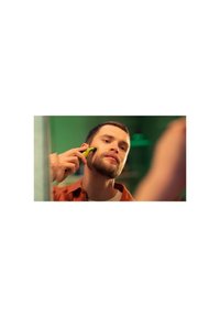 Electric shaver with a bright yellow handle, used by a person with short hair and a beard, in front of a mirror with green background.
