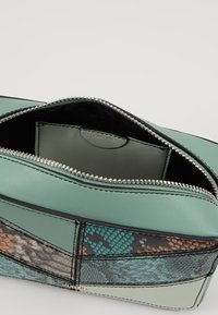 Pochette in pelle patchwork verde e nera con una cerniera argentata visibile parzialmente aperta, che mostra una tasca interna piatta.