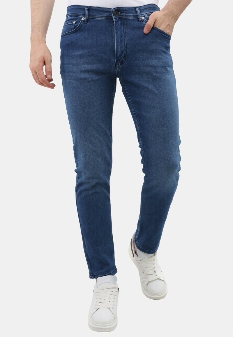 Man in slim-fit blauwe jeans en witte sneakers, met één hand in zijn zak, tegen een effen witte achtergrond.