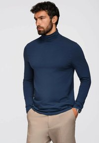 Tummansininen poolopaita, jossa on istuva muotoilu, valmistettu sileästä kankaasta. Pitkät hihat ja ribattu kaulus. Yhdistetty vaaleanruskeisiin housuihin.