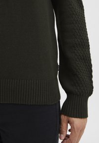 Dunkelgrüner Strickpullover mit strukturiertem Muster an den Ärmeln, gerippten Bündchen und Saum sowie einem dezenten Logosticker an der Seite.