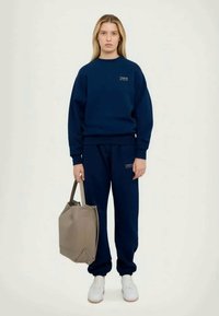 Marinfärgad sweatshirt och byxor i en mjuk bomullsblandning. Sweatshirten har en logotyp på bröstet; modellen håller en ljusgrå tote-väska med handtag.