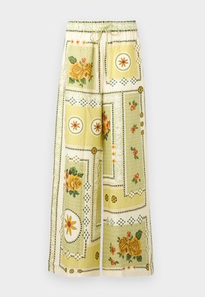 Γυναίκα με μακριά σκουρόχρωμα μαλλιά που φοράει γυαλιά ηλίου, φαρδιά μπλούζα με λουλουδάτο σχέδιο και ταιριαστό παντελόνι φαρδιάς γραμμής, κρατώντας πλεκτή τσάντα, όρθια.