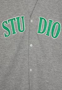 Cardigan grigio con lettere "STUDIO" ricamate in verde. Presenta contorni bianchi e bottoni in plastica sul davanti, mostrando una consistenza morbida.