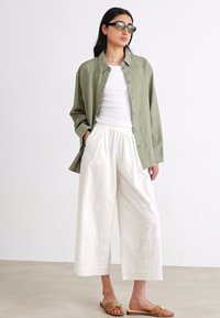 Chemise surdimensionnée verte olive à boutons sur un débardeur blanc, associée à des culottes blanches avec des broderies et des sandales marron clair.