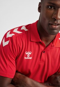 Rode polo shirt van gestructureerde stof, met witte chevrons op de schouders en een logo op de borst. Klassieke kraag en knoopsluiting met drie knopen.