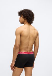 Young man with curly hair wearing black Tommy Hilfiger boxer briefs, standing and facing left showing his bare back against a white background.